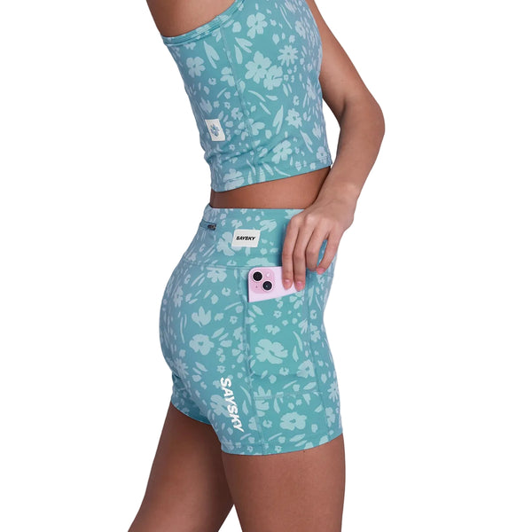 En kvinde i Saysky Flower Combat+ Short Tights 4" Dame, Light Blue Flower, med en matchende crop top, stikker en pink smartphone ned i sidelommen på sine shorts, som har et synligt Saysky-mærke.