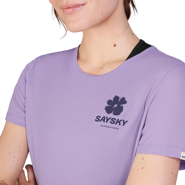 En person er iført en Saysky Flower Flow T-shirt, Dame, Purple, med et sort blomsterlogo på brystet. Kun det nederste af ansigtet, halsen og overkroppen vises mod en hvid baggrund.