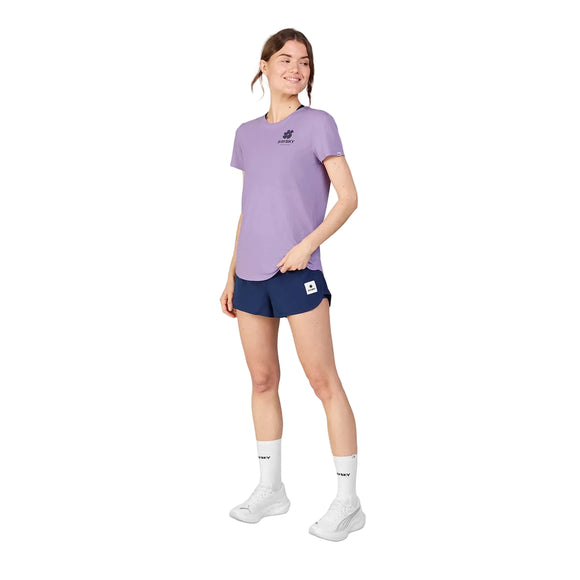 En kvinde smiler iført en Saysky Flower Flow T-shirt, Dame, Purple med marineblå sportsshorts, hvide sokker og hvide sneakers. Hun står let vendt til siden mod en hvid baggrund.