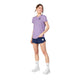 En kvinde smiler iført en Saysky Flower Flow T-shirt, Dame, Purple med marineblå sportsshorts, hvide sokker og hvide sneakers. Hun står let vendt til siden mod en hvid baggrund.