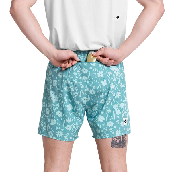 En person står med ryggen til, iført hvid skjorte og Saysky Flower Pace Shorts 5" Herre in Light Blue Flower, og holder et stykke sæbe bag ryggen med begge hænder. En lille tatovering er synlig på bagsiden af det venstre ben.
