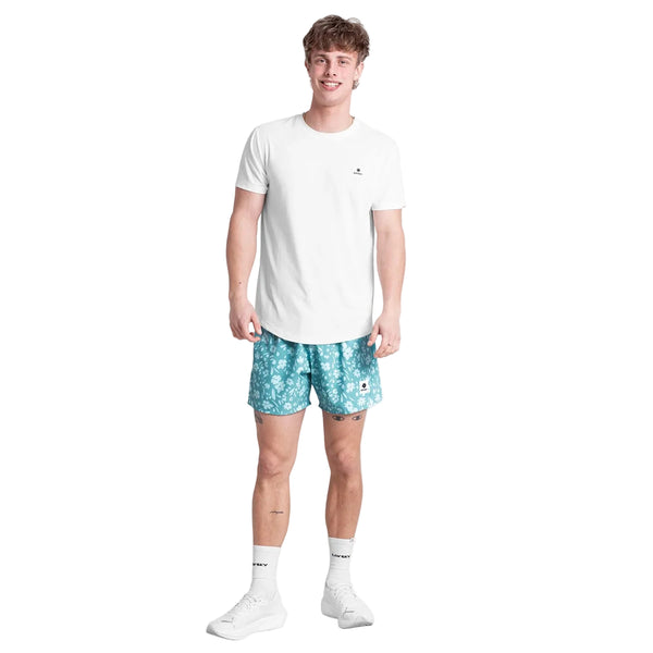 En person står med ansigtet fremad iført en almindelig hvid t-shirt, Saysky Flower Pace Shorts 5" Herre in Light Blue Flower, hvide strømper og hvide sneakers på en hvid baggrund. De lette shorts har et diskret blåt blomstermønster.