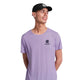 En mand iført Saysky Flower Flow T-shirt, Herre, Purple - med blomstergrafik og logo - står på en hvid baggrund, smiler og kigger til siden.