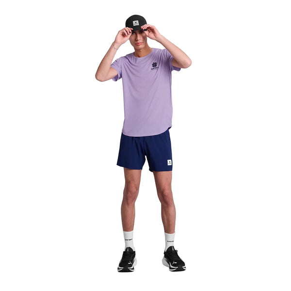 En person står på en hvid baggrund iført en Saysky Flower Flow T-shirt, Herre, Purple, marineblå shorts, hvide sokker, sorte sportssko og en sort baseballkasket, mens han justerer kasketten med begge hænder.