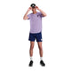 En person står på en hvid baggrund iført en Saysky Flower Flow T-shirt, Herre, Purple, marineblå shorts, hvide sokker, sorte sportssko og en sort baseballkasket, mens han justerer kasketten med begge hænder.