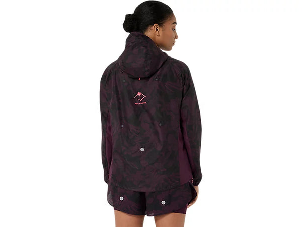 En person med mørkt hår sat op i en knold har Asics Fujitrail Packable-jakken til kvinder i farven Dark Aubergine på, kombineret med matchende shorts. Jakken har et bjergmotiv og et tekstlogo på ryggen. Personen står med ryggen til kameraet.