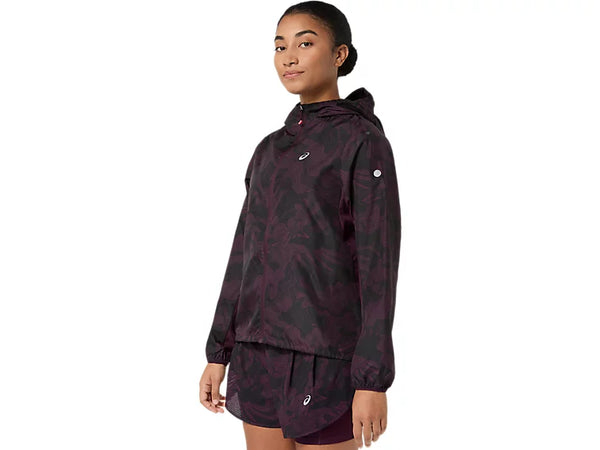 En person står med ansigtet let vendt mod venstre og har en mørk auberginefarvet ASICS Fujitrail Packable-jakke til kvinder på, kombineret med matchende shorts. Tøjet er forsynet med små ASICS-logoer på både jakken og shortsene. Baggrunden er ensfarvet hvid.