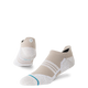 Et par Stance Fusion Grip Tab unisex-sokker i farvevarianten »Croissant«, der har et lavt, sporty design i beige og hvide nuancer, en struktureret sål med Fusion Grip Tab-teknologi, sidestriber, Stance FreshTek-fugtkontrol og et lille cirkulært logo på hælen – vist på en ensfarvet hvid baggrund.