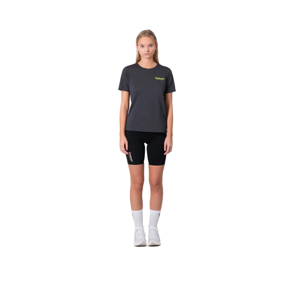 En kvinde står mod en hvid baggrund iført Liiteguard Ground-Tech T-shirt, Dame, i farven Ebony/Neon Yellow. Hun bærer sorte shorts, hvide crew sokker og hvide sneakers med armene afslappet langs siderne - klar til løb.
