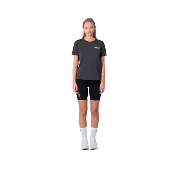 En kvinde står mod en hvid baggrund iført Liiteguard Ground-Tech T-shirt, Dame, i farven Ebony/Neon Yellow. Hun bærer sorte shorts, hvide crew sokker og hvide sneakers med armene afslappet langs siderne - klar til løb.