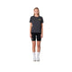 En kvinde står mod en hvid baggrund iført Liiteguard Ground-Tech T-shirt, Dame, i farven Ebony/Neon Yellow. Hun bærer sorte shorts, hvide crew sokker og hvide sneakers med armene afslappet langs siderne - klar til løb.