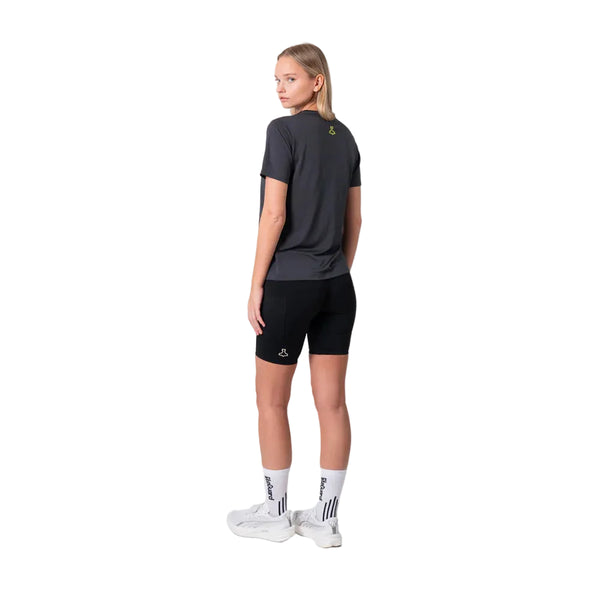 En person står med ryggen til og kigger sig over skulderen. Vedkommende er iført en Liiteguard Ground-Tech T-shirt, Dame, Ebony/Neon Yellow, sorte shorts, hvide strømper og hvide sportssko på en almindelig hvid baggrund.