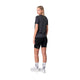 En person står med ryggen til og kigger sig over skulderen. Vedkommende er iført en Liiteguard Ground-Tech T-shirt, Dame, Ebony/Neon Yellow, sorte shorts, hvide strømper og hvide sportssko på en almindelig hvid baggrund.