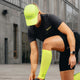 En person i Liiteguard Ground-Tech T-shirt, Dame, Ebony/Neon Yellow, sorte shorts, neonkasket og gule knæstrømper bøjer sig ned for at justere sin strømpe uden for en moderne betonbygning. Vedkommendes ansigt er ikke synligt.