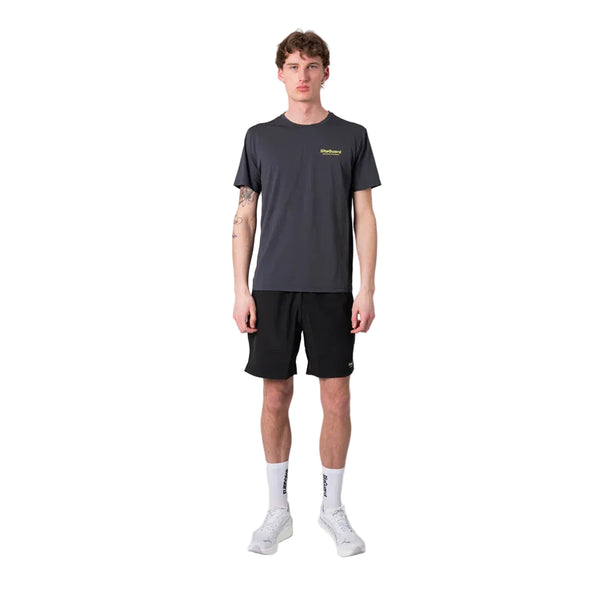En mand står og kigger fremad iført en Liiteguard Ground-Tech T-shirt, Herre, Ebony/Neon Yellow - ideel til løbetræning takket være dens lugtbekæmpende egenskaber - sammen med sorte shorts, hvide sokker og hvide sportssko på en almindelig hvid baggrund.