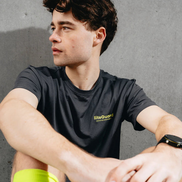 En ung mand med mørkt hår sidder op ad en betonvæg og ser til siden. Han har en Liiteguard Ground-Tech T-shirt, Herre, Ebony/Neon Yellow på, som er lugtbekæmpende og ideel til løb, samt et smartwatch på håndleddet.
