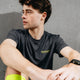 En ung mand med mørkt hår sidder op ad en betonvæg og ser til siden. Han har en Liiteguard Ground-Tech T-shirt, Herre, Ebony/Neon Yellow på, som er lugtbekæmpende og ideel til løb, samt et smartwatch på håndleddet.