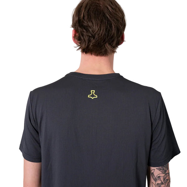 En person med kort brunt hår ses bagfra iført en Liiteguard Ground-Tech T-shirt, Herre, Ebony/Neon Yellow med lugtbekæmpende egenskaber og et lille gult symbol på ryggen. En del af en tatovering kan ses på venstre underarm.