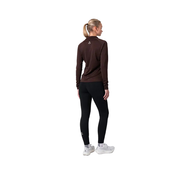 En person står med ryggen til i et simpelt studie iført Liiteguard Ground-Tech 1/4 Zip, Dame, Coffee Bean trøje, sorte leggings, hvide sokker og sneakers. Håret er sat op i en hestehale.