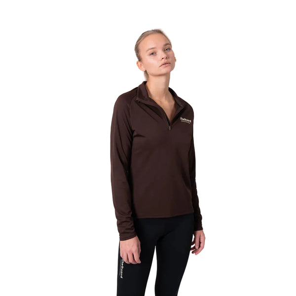 En person iført Liiteguard Ground-Tech 1/4 Zip, Dame i Coffee Bean og sorte bukser står på en almindelig hvid baggrund og ser direkte ind i kameraet med et neutralt udtryk.