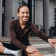 En kvinde i sportstøj og Liiteguard Ground-Tech 1/4 Zip, Dame, Coffee Bean sidder udendørs og strækker benet og smiler, med cykler og en bygning med store vinduer og søjler i baggrunden.