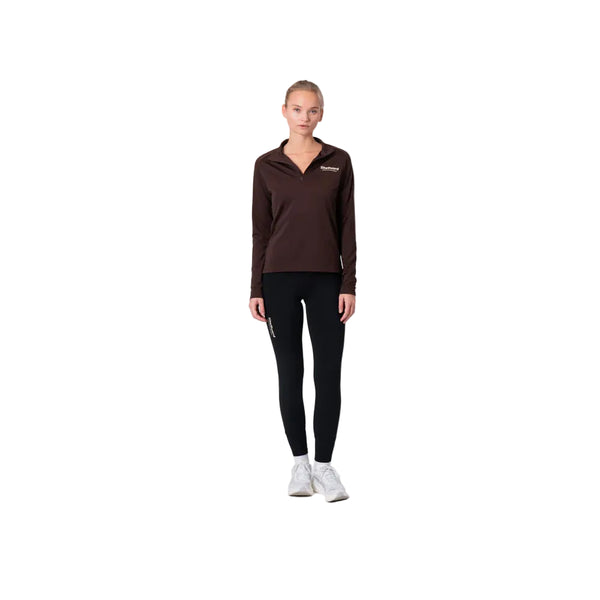 En person står på en almindelig hvid baggrund iført Liiteguard Ground-Tech 1/4 Zip, Dame, i Coffee Bean med et lille logo på brystet sammen med sorte leggings og hvide sneakers. De ser fremad med armene afslappet ned langs siden.
