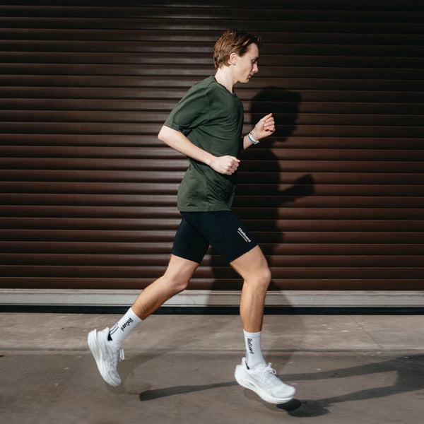 En person i Liiteguard Ground-Tech T-shirt, Herre, Army Green, sorte shorts, hvide sokker og hvide kondisko jogger på en asfalteret overflade foran en brun rulleport.