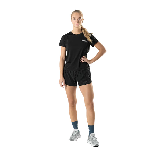 En kvinde kigger fremad mod en hvid baggrund iført Liiteguard Ground-Tech T-shirt, Dame, Black - perfekt til løb - sammen med sorte shorts, blå sokker og grå kondisko. Hendes blonde hår er sat op i en hestehale, og hendes venstre hånd hviler på hoften.