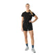 En kvinde kigger fremad mod en hvid baggrund iført Liiteguard Ground-Tech T-shirt, Dame, Black - perfekt til løb - sammen med sorte shorts, blå sokker og grå kondisko. Hendes blonde hår er sat op i en hestehale, og hendes venstre hånd hviler på hoften.