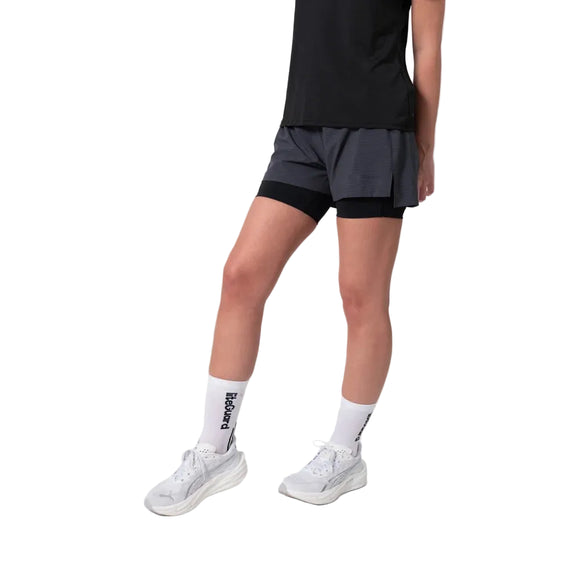 En person iført en sort T-shirt, Liiteguard Glu-Tech Running 2-i-1-shorts til kvinder i farven ebony, hvide sokker med sort tekst og hvide sportssko står foran en hvid baggrund.