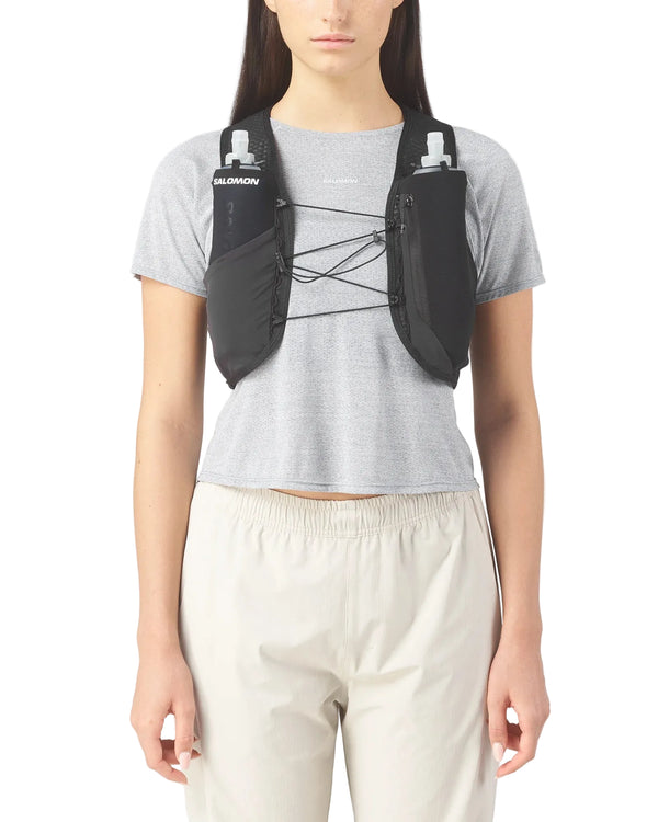 Der vises et billede af en person fra livet og opefter, der står med ansigtet fremad, iført en lysegrå T-shirt med korte ærmer og beige bukser samt et sort Salomon Gravel Skin 4-sæt (unisex) med to synlige vandflaskelommer på brystet.