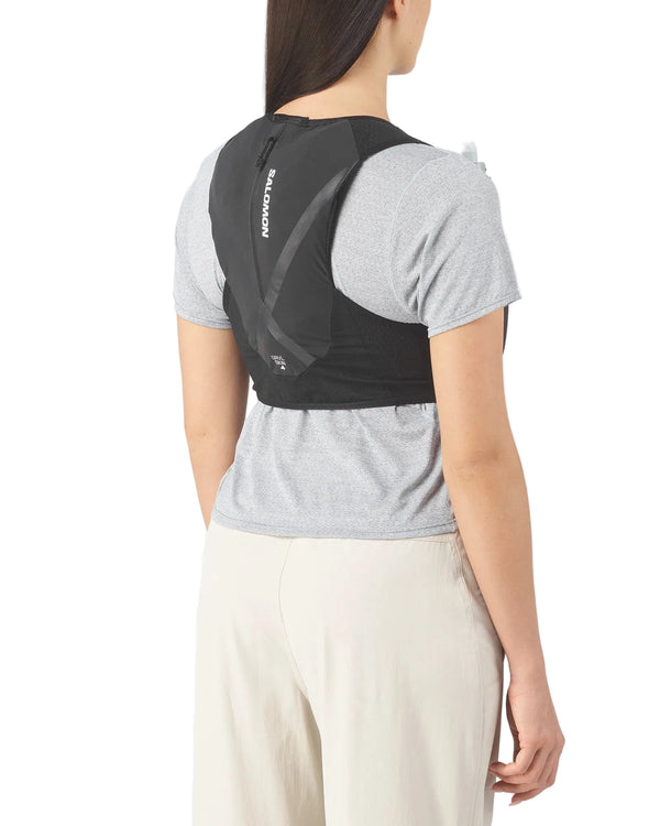 Der ses en person set bagfra, iført en lysegrå T-shirt og beige bukser, som viser den sorte Salomon Gravel Skin 4 Set unisex-rygsæk med synligt logo og tætsiddende design.