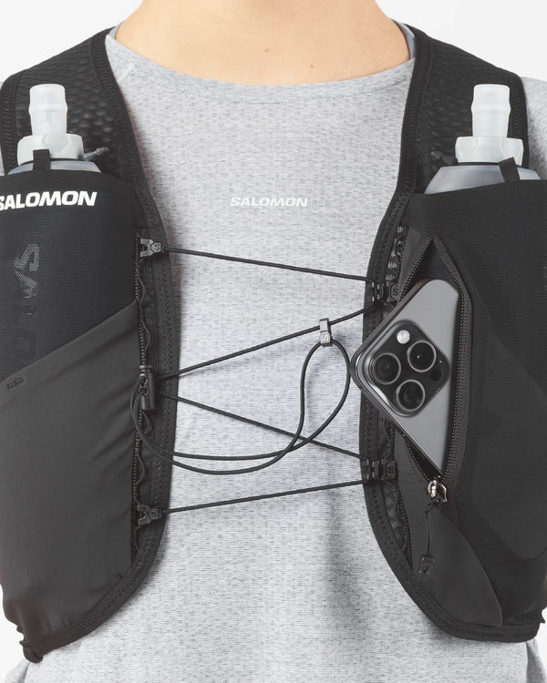 En person iført en grå T-shirt og en sort Salomon Gravel Skin 4 Set løbevest (unisex) med to vandflasker i forlommerne og en smartphone, der delvist kan ses i højre lomme.