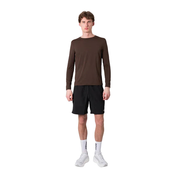 En person står med ansigtet vendt fremad iført en Liiteguard Ground-Tech Light LS T-shirt, Herre, farven Coffee Bean, kombineret med sorte shorts, hvide ankelstrømper og hvide sportssko, foran en ensfarvet hvid baggrund.