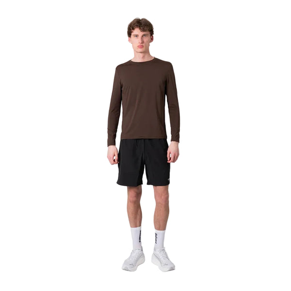 En person står med ansigtet vendt fremad iført en Liiteguard Ground-Tech Light LS T-shirt, Herre, farven Coffee Bean, kombineret med sorte shorts, hvide ankelstrømper og hvide sportssko, foran en ensfarvet hvid baggrund.