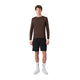 En person står med ansigtet vendt fremad iført en Liiteguard Ground-Tech Light LS T-shirt, Herre, farven Coffee Bean, kombineret med sorte shorts, hvide ankelstrømper og hvide sportssko, foran en ensfarvet hvid baggrund.