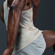 Nærbillede af en person i Nike M Nk Dfadv Aeroswift Jakob Singlet til mænd i Sail/Metallic Gold med Dri-FIT ADV tech og lyse shorts med slidser, vist i bevægelse mod en mørk baggrund; kun en del af overkroppen og benene er synlige.
