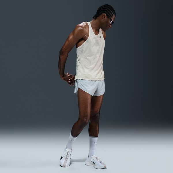 En person iført en Nike M Nk Dfadv Aeroswift Jakob Singlet til mænd i Sail/Metallic Gold, lysegrå løbeshorts, hvide sokker og hvide sportssko står på en almindelig grå baggrund og kigger ned med hænderne foldet bag ryggen.