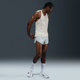 En person iført en Nike M Nk Dfadv Aeroswift Jakob Singlet til mænd i Sail/Metallic Gold, lysegrå løbeshorts, hvide sokker og hvide sportssko står på en almindelig grå baggrund og kigger ned med hænderne foldet bag ryggen.