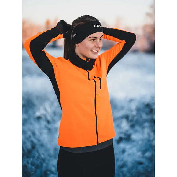 En person i orange jakke og Fusion Headband, Unisex, Black står udendørs i et snedækket landskab, binder håret tilbage og smiler under vintertræningen.