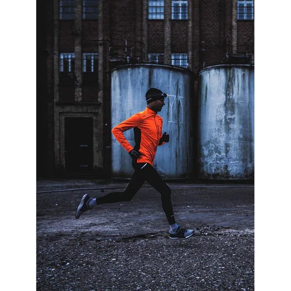 En person løber udendørs på grus, forbi industritanke og en murstensbygning, iført Fusion's Hot Long Tights Unisex i sort.