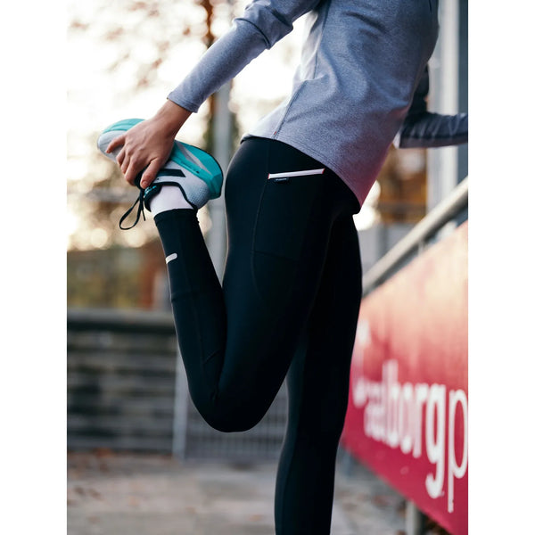 En person i Fusion Hot X-Long Tights (Unisex, sort), en lysegrå langærmet top og blågrønne løbesko strækker benet ved at holde foden bag sig i nærheden af et rødt rækværk udendørs.