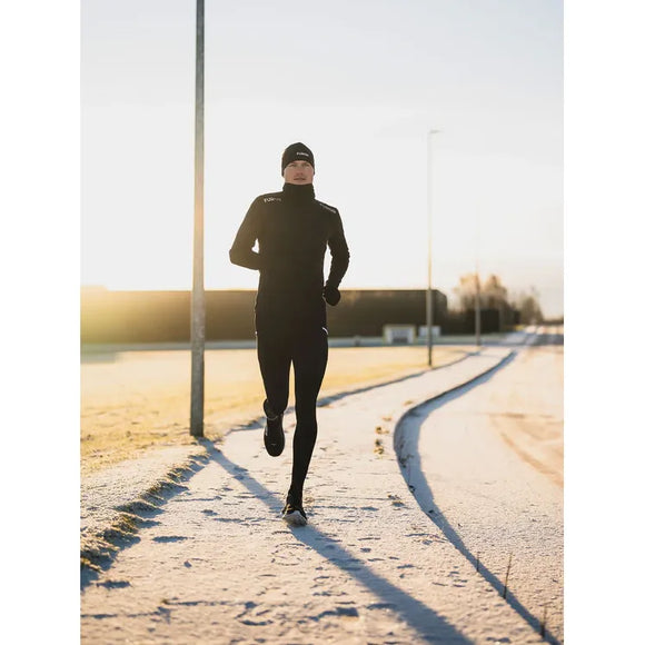 Iført Fusion's Hot X-Long Tights i sort jogger en person på et snedækket fortov ved solopgang eller solnedgang med frostklart græs og bygninger i baggrunden.