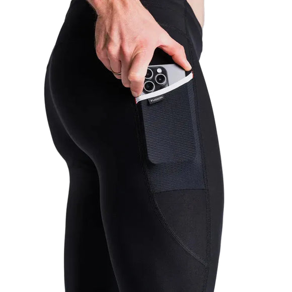En person i Fusion Hot X-Long Tights, Unisex Black, placerer en smartphone med multiobjektiv i sidelommen på leggingsene mod en almindelig lys baggrund.