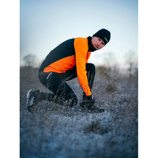 En person klædt i Fusion Hot X-Long Tights (Unisex, sort) knæler på frossent græs og justerer sin sko i et sløret, vinterligt miljø.