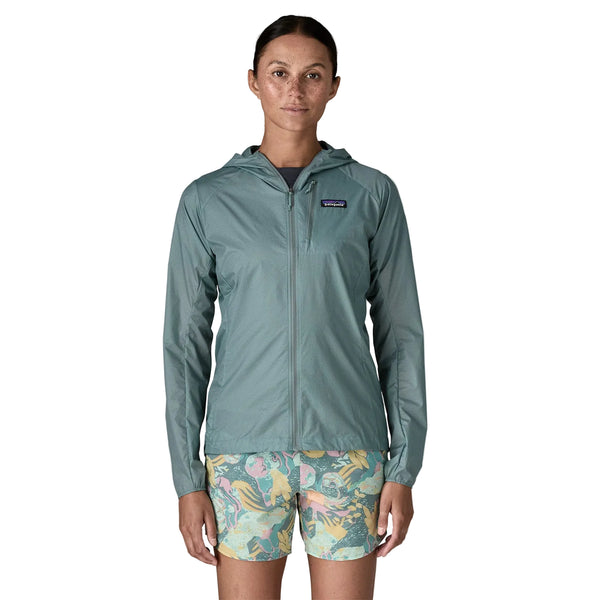 En person står med ansigtet fremad i en Patagonia Houdini Jacket, Dame, Blue Sage - en populær letvægtsjakke dame - med et lille logo på venstre bryst, parret med blå, gule og pink mønstrede shorts. Baggrunden er helt hvid.