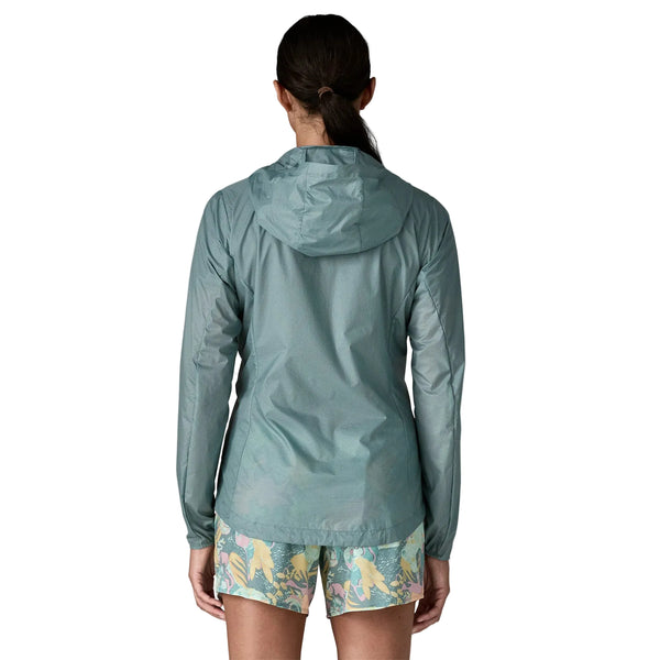 En person står med ryggen til kameraet iført en Patagonia Houdini Jacket, Dame in Blue Sage og shorts med botanisk print på en almindelig hvid baggrund.