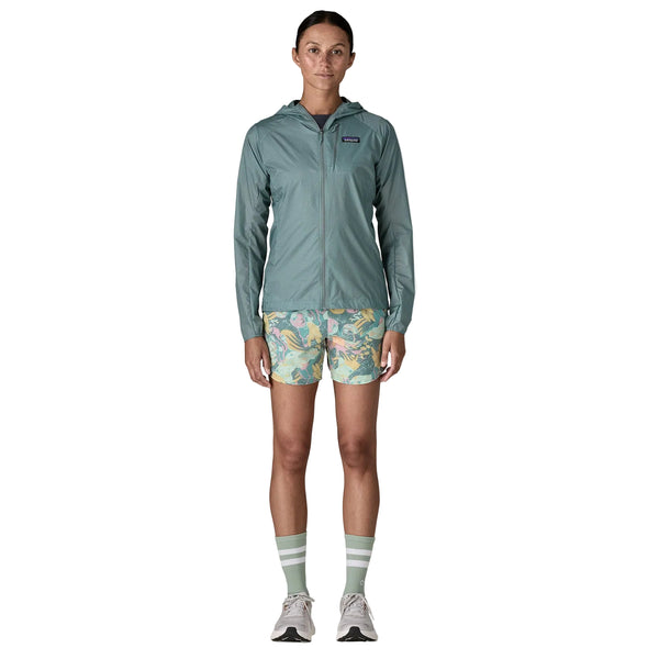 En person står med ansigtet fremad i en Patagonia Houdini Jacket Dame in Blue Sage, mønstrede grønne og gule shorts, stribede sokker og lyse sneakers på en hvid baggrund.