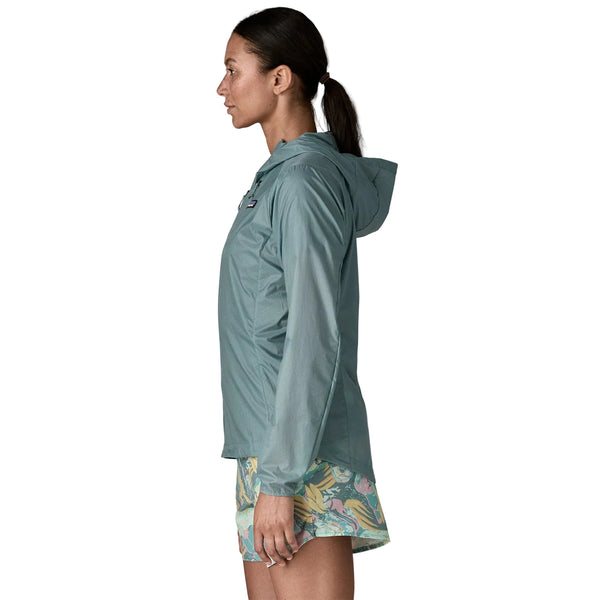 En kvinde står i profil iført en Patagonia Houdini Jacket, Dame, Blue Sage, en letvægtsjakke til kvinder, parret med mønstrede shorts på en hvid baggrund.