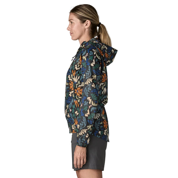 En kvinde i profil bærer en Patagonia Houdini Jacket, Dame, Kaleido: Sort med blå, grønne, orange og beige mønstre. Hendes hår er sat op i en hestehale, mens hun ser til venstre mod en ensfarvet baggrund.